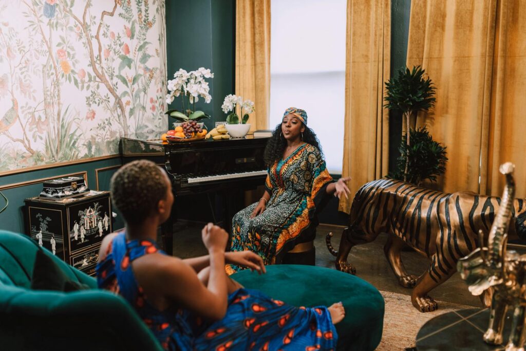 Two women in traditional clothing, with a piano and exotic decor, indoors.