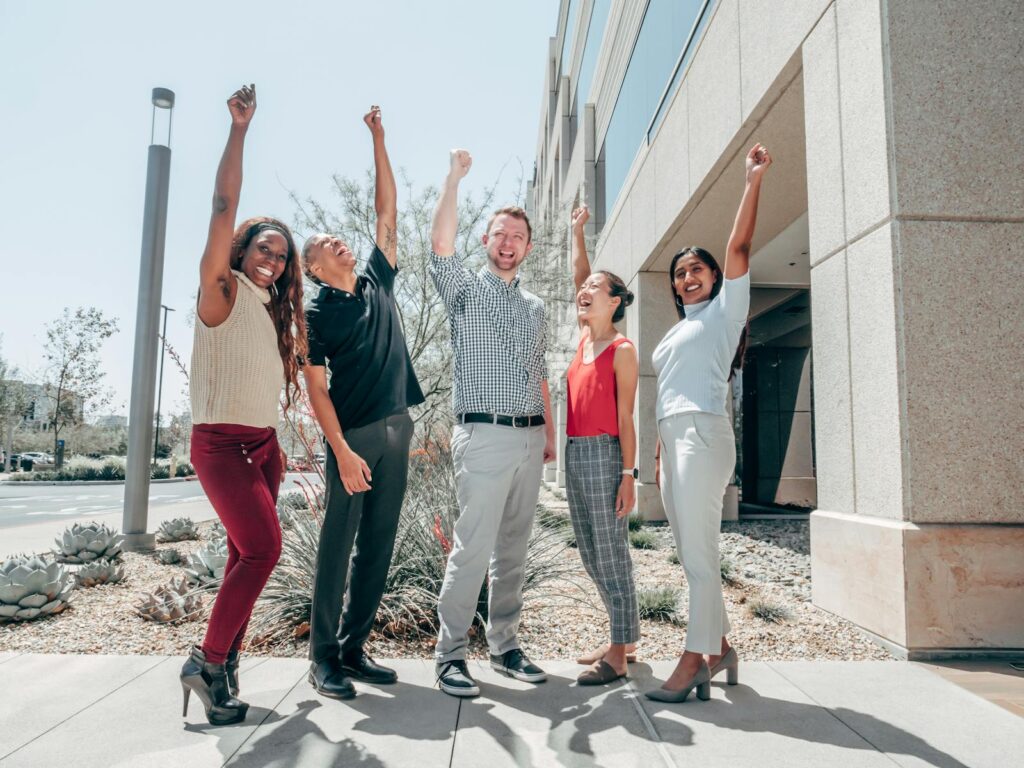 A diverse group of coworkers celebrating excitement and teamwork in a sunny outdoor office setting.