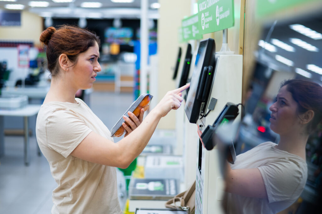 A Frustrated Woman Uses A Self Checkout Counter. The Girl Does Not Understand How To Independently Buy Groceries In The Supermarket Without A Seller