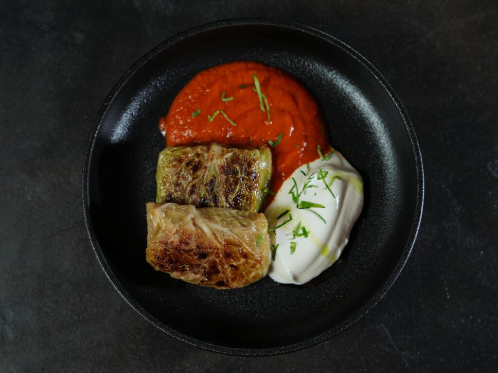 A scrumptious serving of cabbage rolls with tomato and yogurt sauces on a black plate.