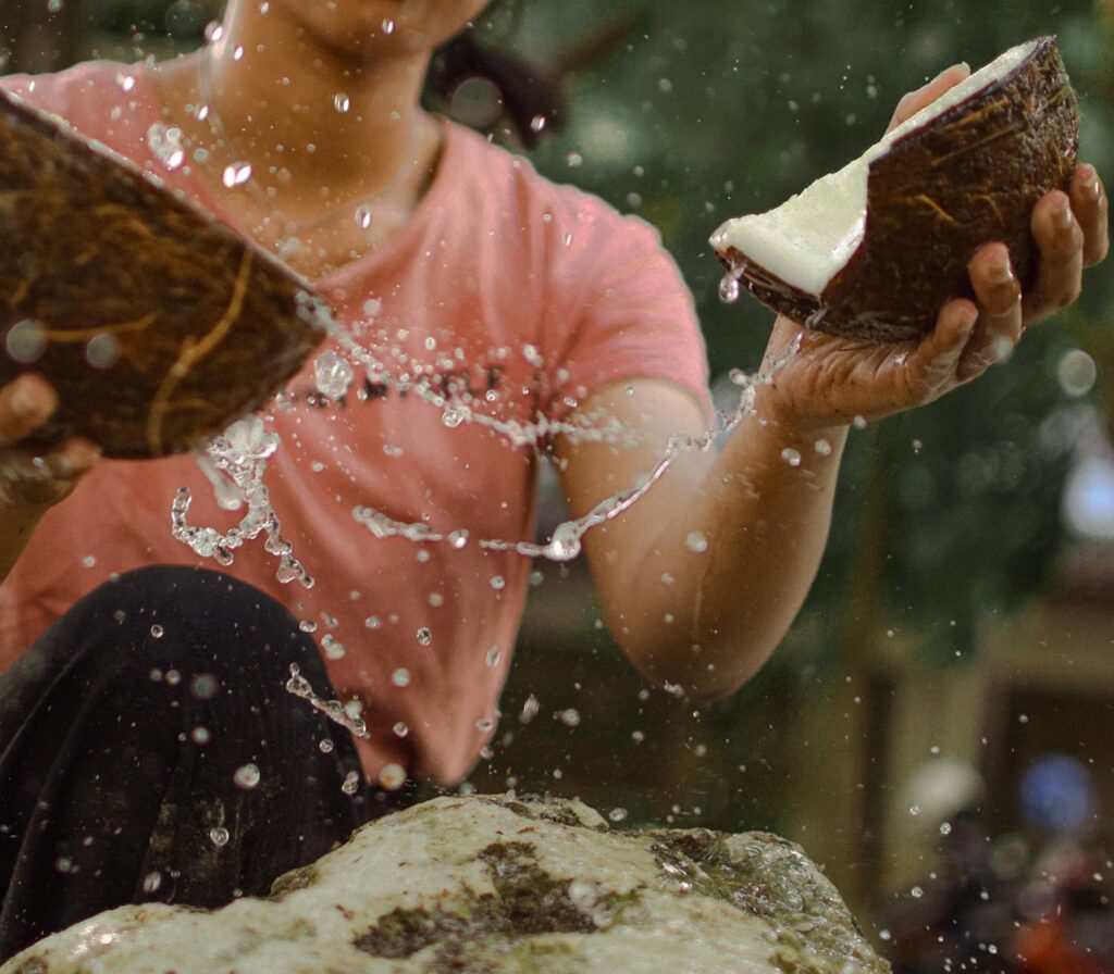 Close-up of hands breaking a coconut, with refreshing juice splashing outdoors.
