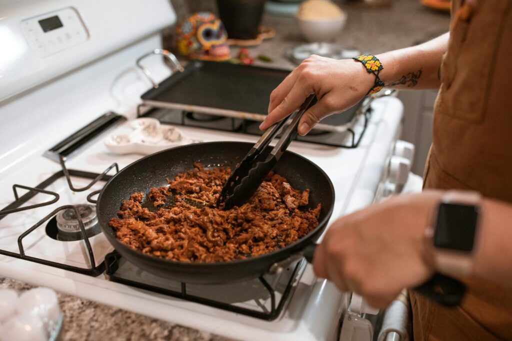 Hands cooking taco meat in a pan on a gas stove with tongs.