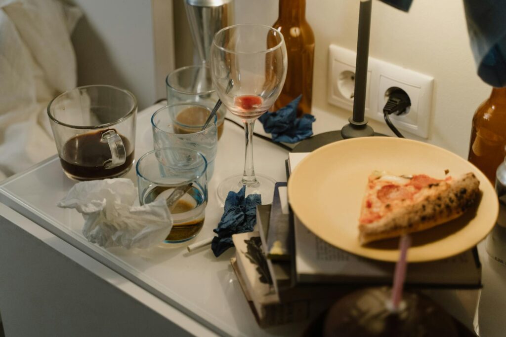 A cluttered nightstand filled with dirty glasses, tissues, and a pizza slice on a plate.
