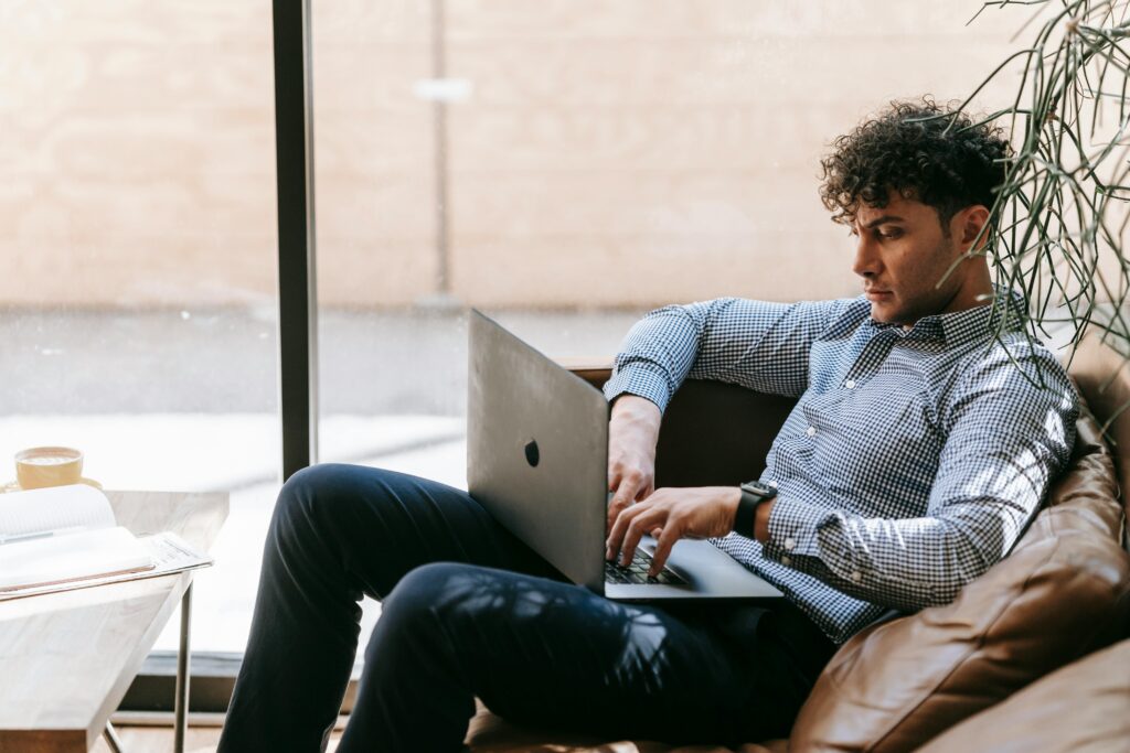 A man lounges on a couch, eyes glued to a laptop screen, while a dusty treadmill sits nearby