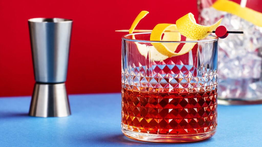 Sazerac, Alcoholic Cocktail Drink With Cognac, Bourbon, Absinthe, Bitters, Sugar And Lemon Zest. Dazzling Red Blue Background With Hard Light And Shadows Pattern