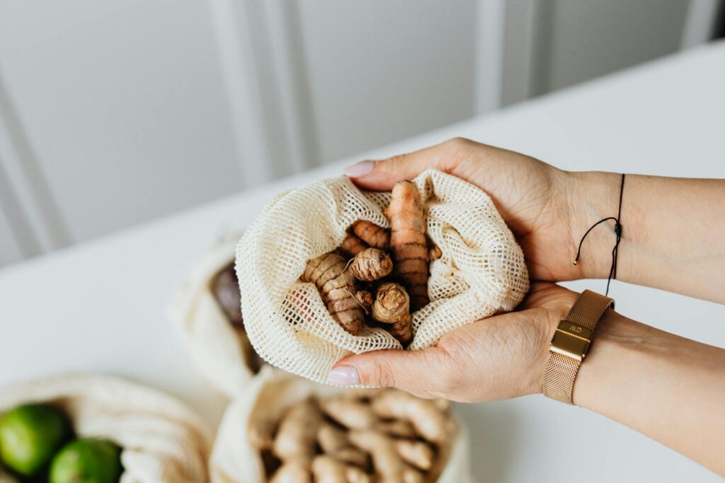 Hands holding fresh turmeric in a mesh bag on a white table, highlighting natural ingredients.