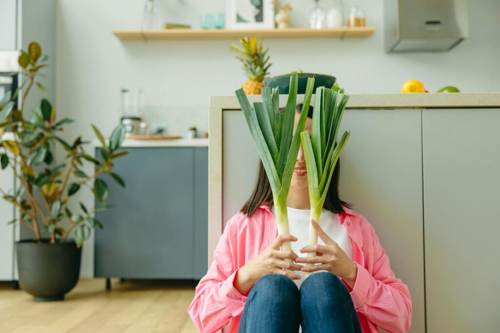 A woman in a pink shirt playfully hides behind leeks in a modern kitchen setting.
